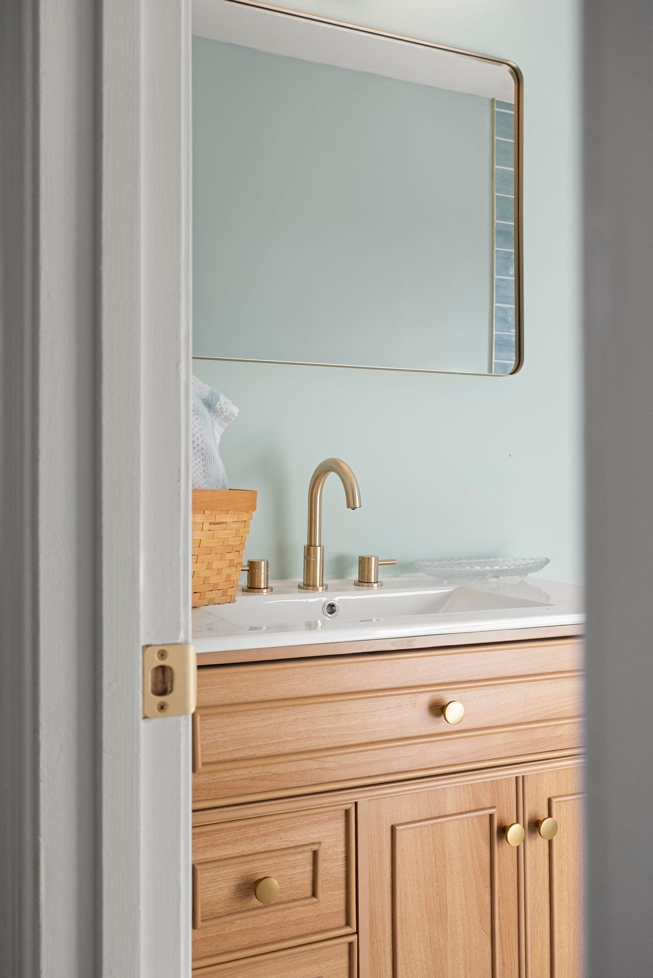 Bathroom with a natural wood vanity, brass widespread faucet and rounded mirror on a pale green wall