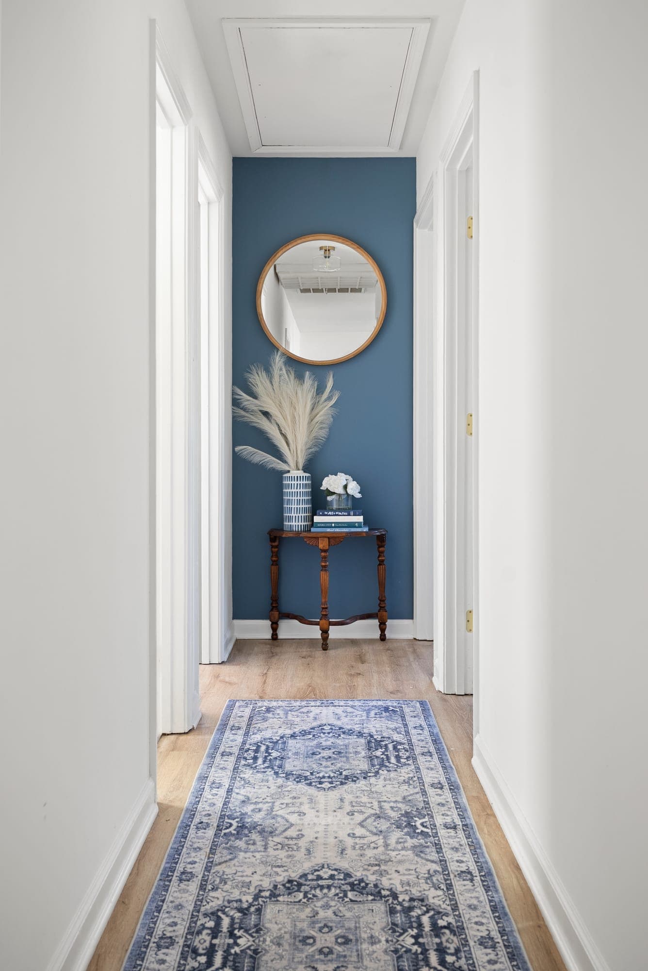 Hallway with a deep blue painted accent wall, round mirror and patterned runner on plank flooring