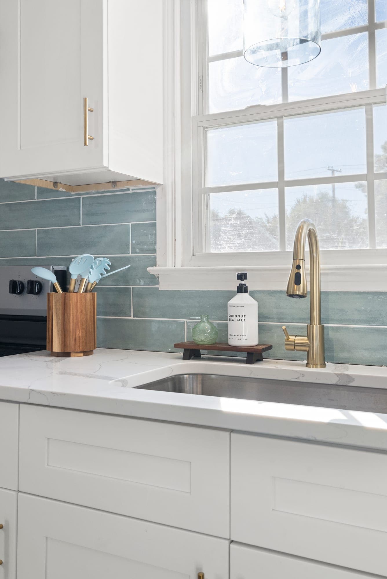 Kitchen sink under a window with a brass pull-down faucet, quartz countertop and tile backsplash